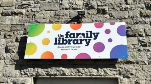 A stone wall with a sign which reads "The Family Library: books, activities and so much more!" It is white with multi-coloured spots.
