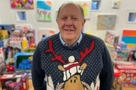 Martin Crew standing in a room full of toys and games smiling at the camera. He has a bald head with grey hair at the sides. He is wearing a blue Christmas jumper with white "snow" on it along with a big reindeer's face. The reindeer has red antlers and is wearing a white, red and black bobble hat.