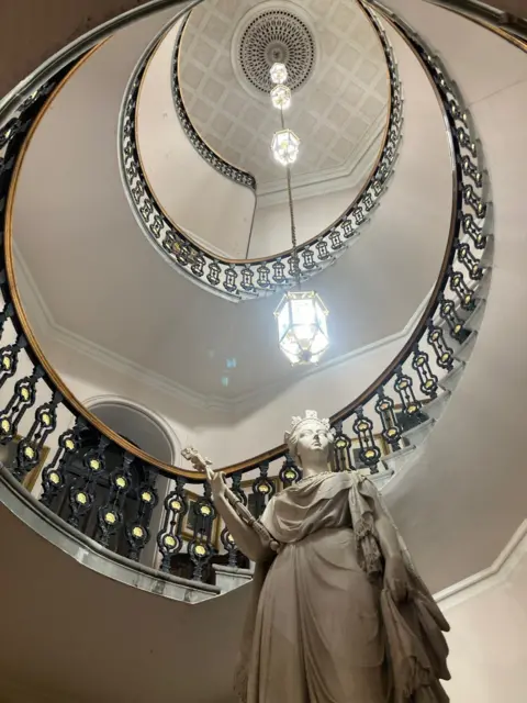 Andrew Maywood A marble statue of Queen Victoria, photographed from below, with a winding staircase descending up and an ornate light hanging from the ceiling