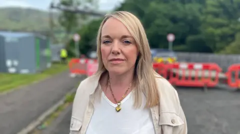BBC Ellen Forson, standing on a residential street in front of red safety barriers that block off the road, with trees and a hillside in the background. She is wearing a light-coloured jacket over a white top and a chain necklace, and is positioned in the foreground with the background softly out of focus.