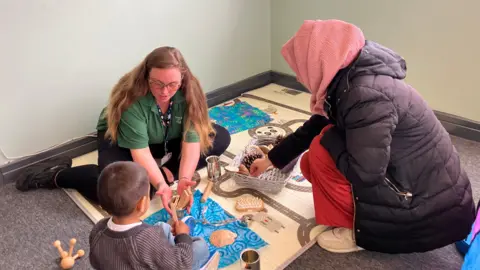 Shariqua Ahmed/BBC A woman in a red head-scarf and black jacket, playing with a boy in a grey jumper and a woman in a green T-shirt showing them toys.