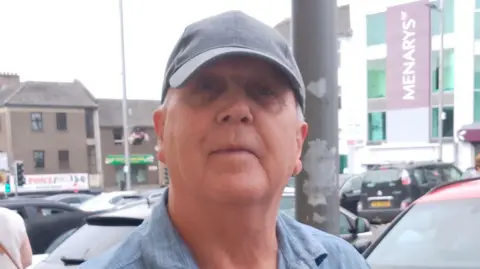 Kevin is wearing a grey cap and a blue shirt. He's standing in front of Menarys shopping centre.