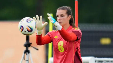 Olive Clark, with brown hair in a ponytail, has her arms out in front of her. She is about to catch a ball which is travelling towards her. She is wearing a red football shirt with EE in yellow letters. She is wearing white goalkeeper gloves. 
