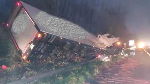 A white lorry hanging off the side of a motorway. It is still dark.