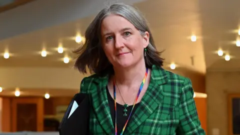 Maree Tood has shoulder length grey hair and is wearing a green tartan jacket over a black top. She is walking through a corridor in the Scottish Parliament.
