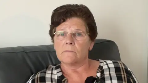 Joan is sitting on her grey sofa and looking slightly off to the right of the camera lense. She is where a grey, white and black check shirt. She has short dark hair, blue eyes and is wearing silver rimmed glasses.
