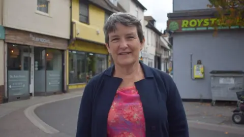 Ruth Brown A picture of Councillor Ruth Brown who is a woman with short greyish black hair. She is wearing a dark blue jacket and a blouse that is pink but with patches of blue, red and yellow on it. She is stood in the streets in Royston, with a beauty salon behind her in a pedestrian part of the town.