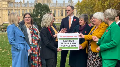 Qays Najm/BBC MPs and campaigners stand outside parliament holding a box which says "Save our Arthur Rank Hospice Beds".