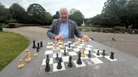 Graham Chesters has white hair and is wearing a grey jacket with a blue shirt underneath. He is sat down at a concrete chess board in a park near a pond. The black and white chess pieces are scattered around the board. He has just achieved checkmate with his queen and bishop angled towards the opposing black king.