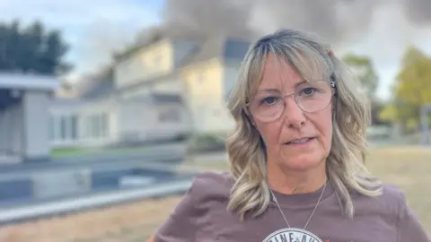Stephen Huntley/BBC A woman with blonde hair and glasses looks into the camera. She is standing in front of a large house and pool. Behind the house large plumes of grey smoke are going into the air.