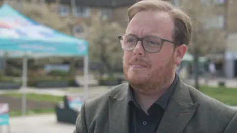 Man wearing glasses and a dark blazer standing outdoors, with a campaign-style gazebo and signage blurred in the background on a public green