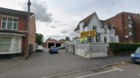 Google A white building is near the centre of the photo with a yellow sign, with words including hand car wash in black on it, close to the building. Other buildings are on the far left and right of the image.