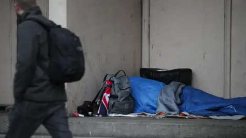 A homeless person sleeping rough in a blue sleeping bag a doorway. A man in a black coat, black backpack and dark blue jeans walks past.