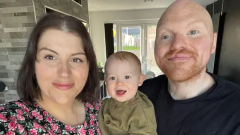 A woman with black hair and a pink flowery top and a bald man with a ginger beard. Between then they are holding a ginger baby in a green jumper.