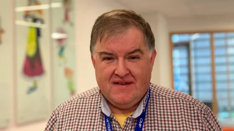 A man with brown hair is looking directly at the camera. He is smiling. He is wearing a check shirt and a blue string around his neck. he is in a hospital setting