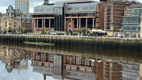 Newcastle Crown Court reflected in the River Tyne running in front of it. It is an imposing building made from smooth red stone with massive black windows and tall columns along its frontage.