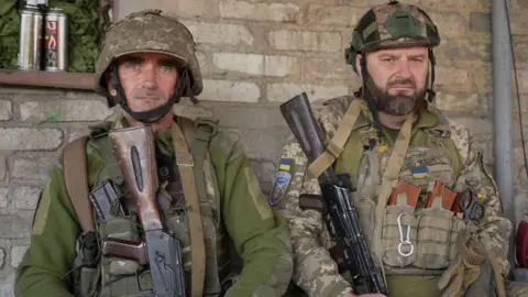  Two Ukrainian soldiers wearing helmets and carrying guns look into the camera lens