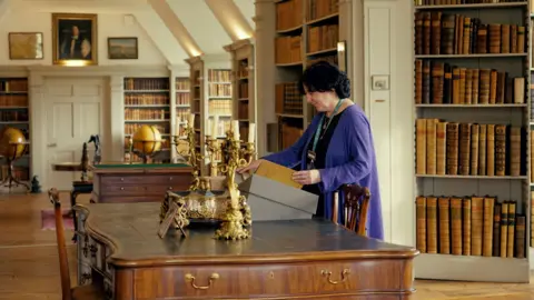 A view of a large room with bookcases on almost every wall filled with old documents and manuscripts. A woman in the centre is looking at a book on a large mahogany table.