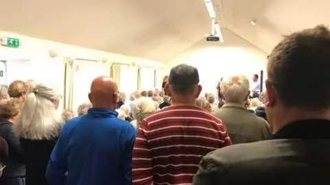 Karen Froggatt A village hall full of people. In front there are the backs of three men who are standing in front of them a lot of people are sitting down. 