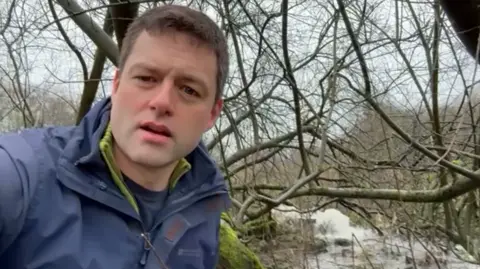 A clean-shaven man with brown, short cropped hair speaks with a river behind him.