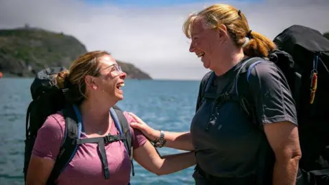 BBC / Studio Lambert Cathie and Tricia pictured facing one another smiling. They are holding each others arms and wearing backpacks. 