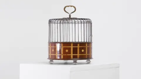 Sworders A wooden birdcage placed on top of a white table. The birdcage is brown and has a round handle on top.