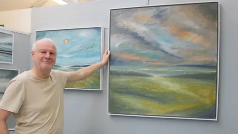 Lansdown Art Studios Association Tony, smiling to camera, and holding up a large painting of a landscape of green fields, and blue and orange sky