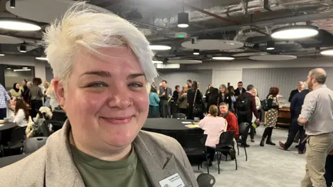 Amy Holmes/BBC A woman wearing a light brown blazer with a pink pin and a green top stands in the foreground of a busy event space. Behind them, groups of people are gathered around tables, chatting and moving around the large room. The venue has exposed ceiling fixtures, circular lights, and modern grey décor, with a lively atmosphere as attendees network and socialise.
