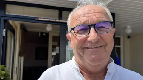 A white man standing outside in front of a building with an open door behind him. He is dressed in a white shirt with a tie detail at the collar. There is visible greenery, while the building’s roof and walls frame the background. The sky above is clear and blue, suggesting a bright, sunny day.