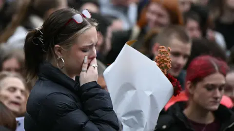 Getty A young woman wearing a black jacket holds a bouquet of flowers, one hand covering her mouth as she looks down, at a vigil with a crowd in the background