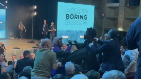 Hecklers in the audience being tackled by other men, with the stage in the background showing a screen with the words 'Boring, Boring, Boring' written on it.