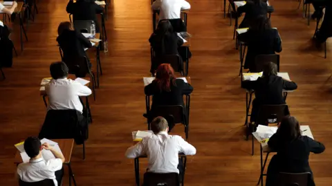Rows of pupils doing exams in a hall. Some are wearing blazers while others have white shirts on. There is a mix of girls and boys and all have their heads down and are facing their work so we can only see the backs of their heads.