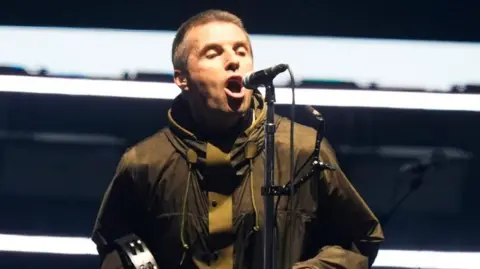 PA Media Liam Gallagher stands on stage singing into a microphone and playing a tambourine.