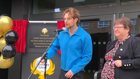 Central Bedfordshire Tom Grennan pictured behind a microphone at Sandy Secondary School. He wears a bright blue long sleeve shirt with black buttons. He has curly hair, a gold ear piercing and a tattoo on his neck. On his right is Karen Hayward who has short brown hair, a black blazer with gold buttons and is wearing a floral dress. 