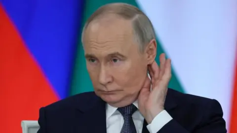 Pictured in front of the colours of the Russian flag, Putin, wearing a suit, looks to the left of the camera, pressing two fingers behind his ear