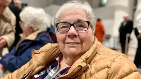 Katrina Hughes is smiling at the camera. She has cropped white hair and black-rimmed glasses and is wearing a camel coloured down coat with a plaid shirt underneath. In the background other people can be seen seated at the public meeting about Brynmawr Medical Practice. 