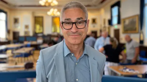 Gerald Voisin is wearing a pale blue-grey open neck shirt and jacket and is standing in a large hall with tables and paintings in the background.