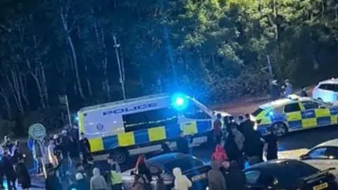 A white, blue and yellow police van and a police car are parked in front of a crowd of people and various cars at an evening gathering