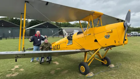 Care UK Two men, with their arms out out, by a yellow plane. Doug, in a wheelchair, to the right, is wearing a green coat, with a hat on, with short grey hair, the other man, David, has on a lanyard, blue jeans and a blue top. They are both smiling, with buildings behind them.