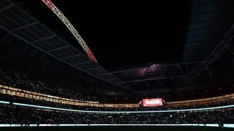 Wembley Stadium dark with tiny lights held by people in the stands watching the match