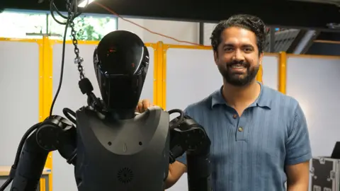 Zoe Corbyn Wearing a blue t-shirt, Sankaet Pathak stands next to the black Phantom robot.