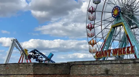 Fairground rides, including a ferris wheel, can be seen over a brick wall at Skegness Pleasure Beach. You can just see the people who are stuck on the ride.