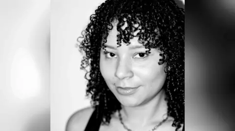 Andrezza Vieria A black and white picture of Mali looking at the camera with a toothless smile. she has shoulder length curly black hair with a fringe