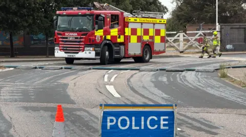Richard Knights/BBC A nearby road to a warehouse which is on fire. The road has been closed by police and within the cordon is a fire engine. Two fire fighters are walking towards the back of the fire engine which is parked on the road.