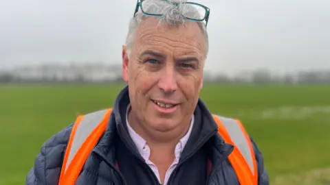 A man wearing an open-collared shirt, jacket, coat and orange high-vis looking at the camera. He has glasses and grey hair, and is standing in a field.