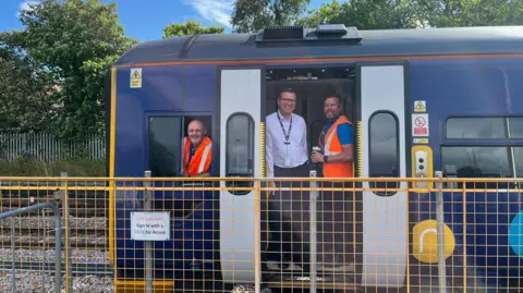 Northumberland County Council A Northern train on the Northumberland Line. A drive is peering out of the window. A conductors and member of staff are stood in the door.