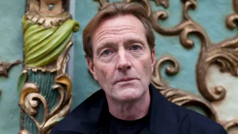 A man with brown hair and a navy coat looks into the camera with a neutral expression