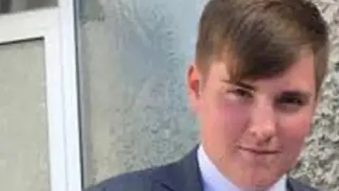 RTÉ NEWS Cameron Reilly. He has short light brown hair, wearing a dark grey blazer and white shirt.