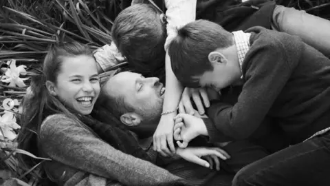 A black and white photo of Prince William lying on the grass - his three children hug him while Charlotte looks at the camera and smiles