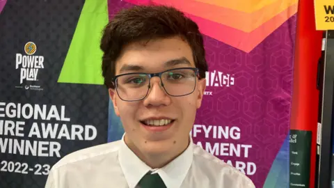 Jonathan smiling at the camera. He is wearing glasses, a white shirt and green tie. He has dark brown hair.